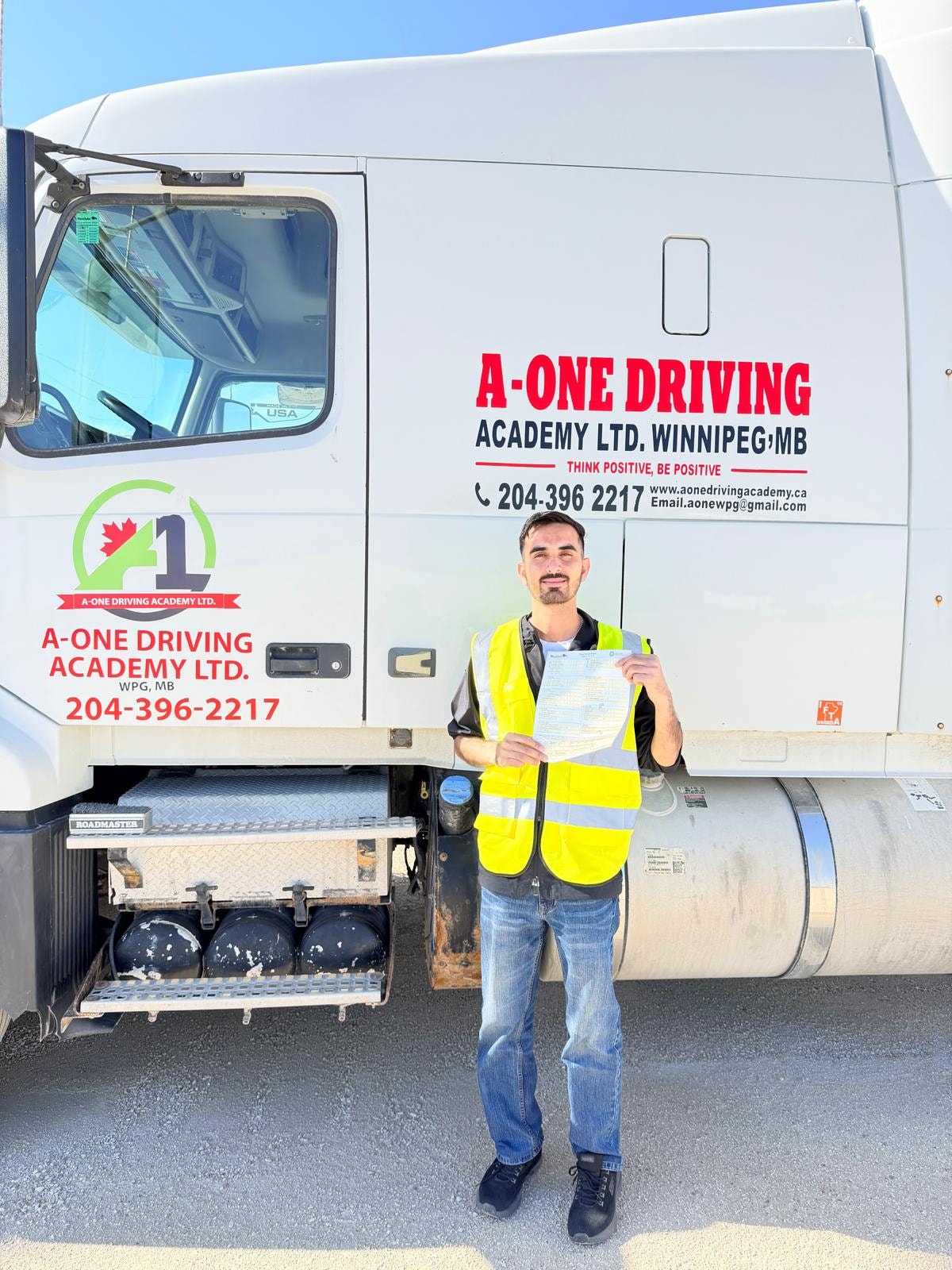 Student in yellow safety vest holding road test pass document in front of A-One Driving Academy truck